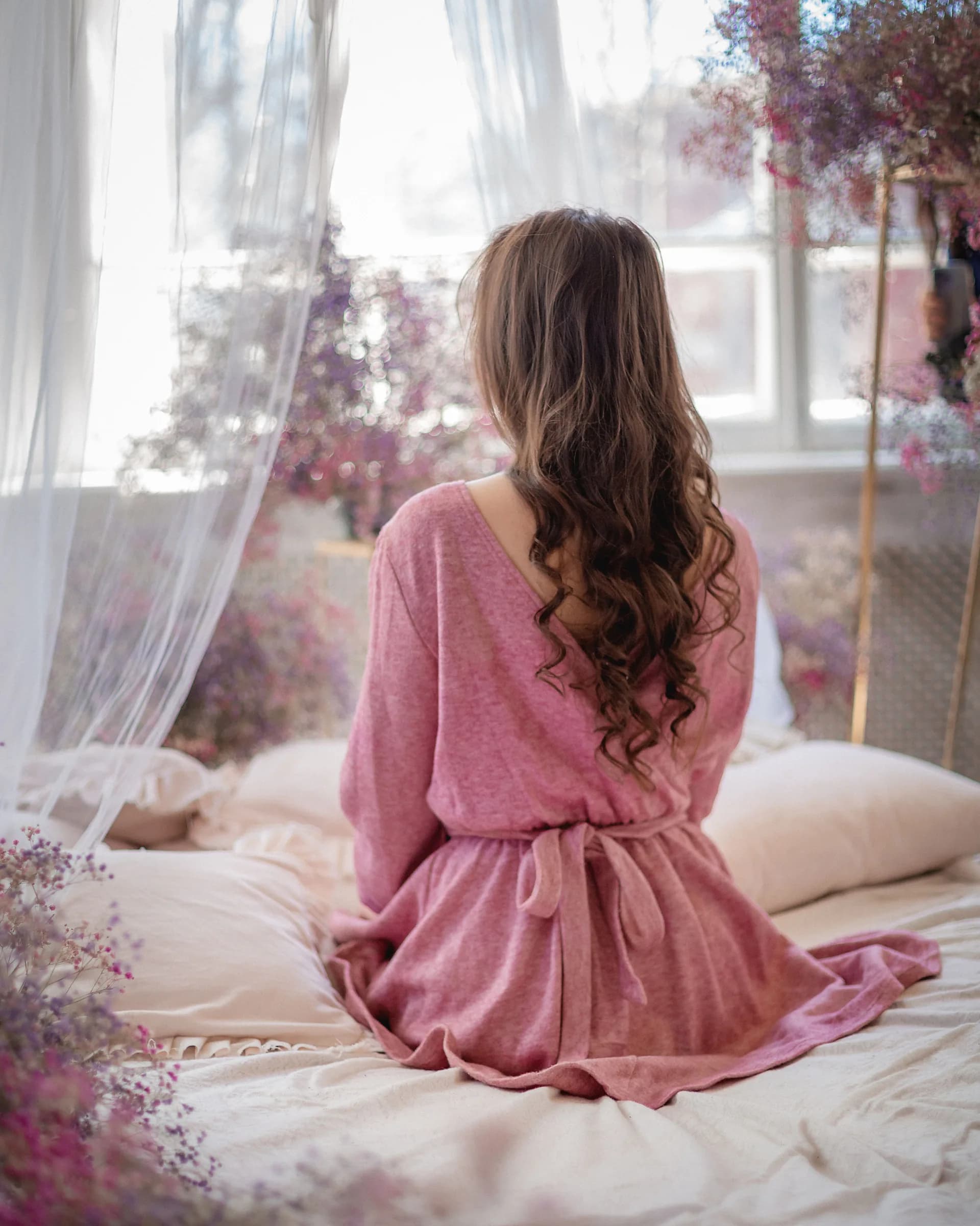 A woman in a soft pink robe sits on a bed by a sunlit window, framed by sheer curtains and pink blossom